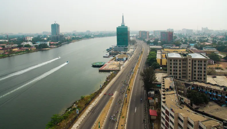 Vue aérienne d'un boulevard urbain longeant un fleuve, avec un gratte-ciel en verre, un héliport et un hors-bord.
